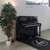 Un piano droit noir Fridolin F121 est placé à côté d'une plante en pot, dans une pièce moderne au sol en béton.