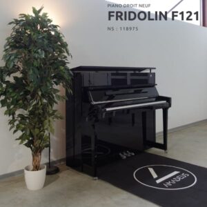 Un piano droit noir Fridolin F121 est placé à côté d'une plante en pot, dans une pièce moderne au sol en béton.