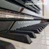 Un piano droit neuf Fridolin F121 de Schimmel, photographié en gros plan, montrant les touches noires et blanches et des détails métalliques brillants.