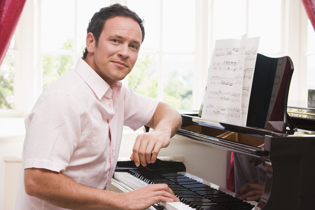 Homme jouant du piano devant une fenêtre lumineuse avec une partition, portant une chemise blanche, souriant légèrement.