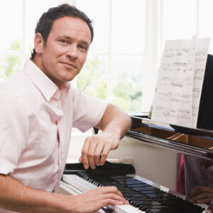 Homme jouant du piano devant une fenêtre lumineuse avec une partition, portant une chemise blanche, souriant légèrement.