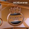 Le logo du piano Wilhelm Schimmel W180 en bronze, avec des détails gravés et une couronne en relief stylisée. Le piano est un quart de queue.