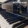 Gros plan d'un piano à queue Wilhelm Schimmel W180 dans une salle de musique bien éclairée, avec plusieurs autres pianos en arrière-plan.