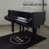 Un piano à queue noir "WILHELM W180" est posé sur un tapis noir, sur un sol gris dans une pièce.