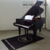 Un piano à queue Wilhelm W180 noir est exposé dans une pièce, avec une couverture noire en dessous portant un texte.