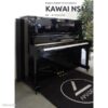 Piano droit d'occasion Kawai NS15 en noir laqué, numéroté K1565293, exposé sur un tapis noir près d'une plante verte.