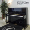 Piano droit d'occasion Yamaha U1 noir brillant avec numéro de série 4933077, exposé dans un showroom avec plante et lampe.