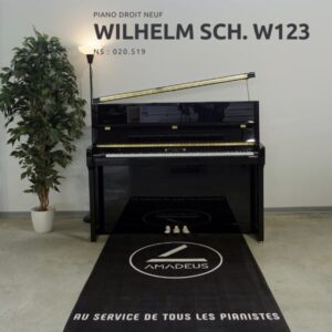 Un piano droit noir Wilhelm Sch. W123 est exposé sur un tapis Amadeus, accompagné d'une lampe et d'une plante verte en pot.